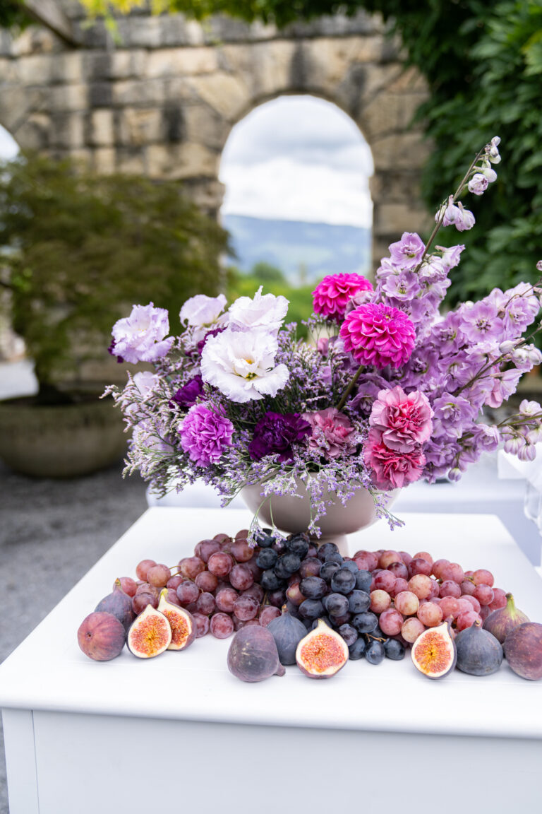 Ein Strauß aus rosafarbenen und violetten Blumen mit Weintrauben und Feigen auf einem weißen Tisch im Freien - die perfekte Inspiration für Hochzeitsplaner in Italien, die auf der Suche nach einzigartigen Akzenten für die Hochzeitsplanung sind.