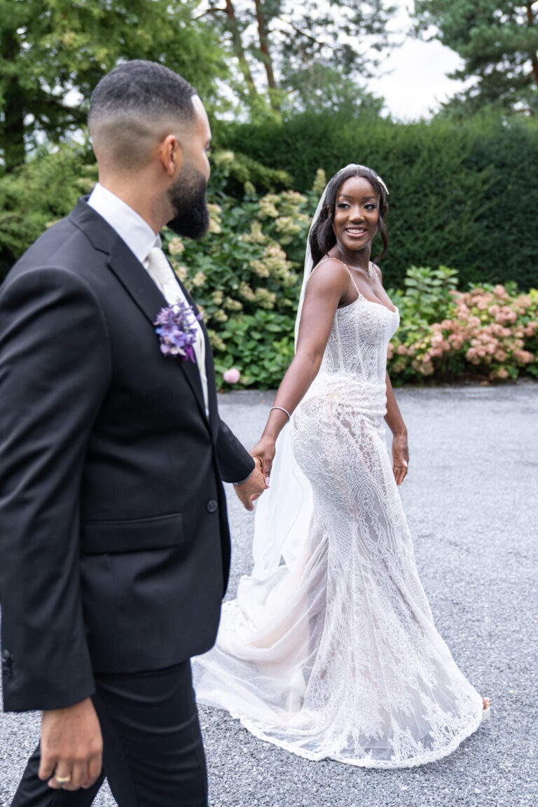 Eine Braut im Spitzenkleid lächelt, während sie mit ihrem Bräutigam im schwarzen Anzug unter freiem Himmel Händchen hält, während ihr Hochzeitsplaner dafür sorgt, dass jedes Detail ihrer italienischen Feier einfach perfekt ist.
