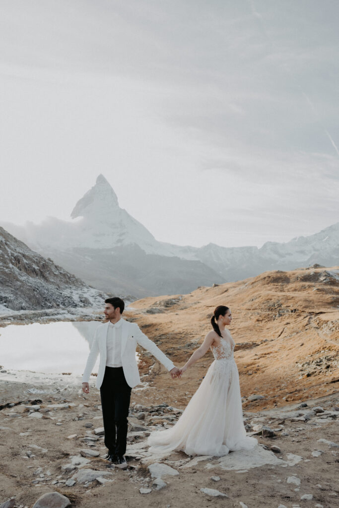 Ein Paar in Hochzeitskleidung hält sich in einer atemberaubenden Berglandschaft mit einem schneebedeckten Gipfel im Hintergrund an den Händen - die perfekte Inspiration für alle Hochzeitsplaner, die atemberaubende italienische Hochzeitsziele suchen.