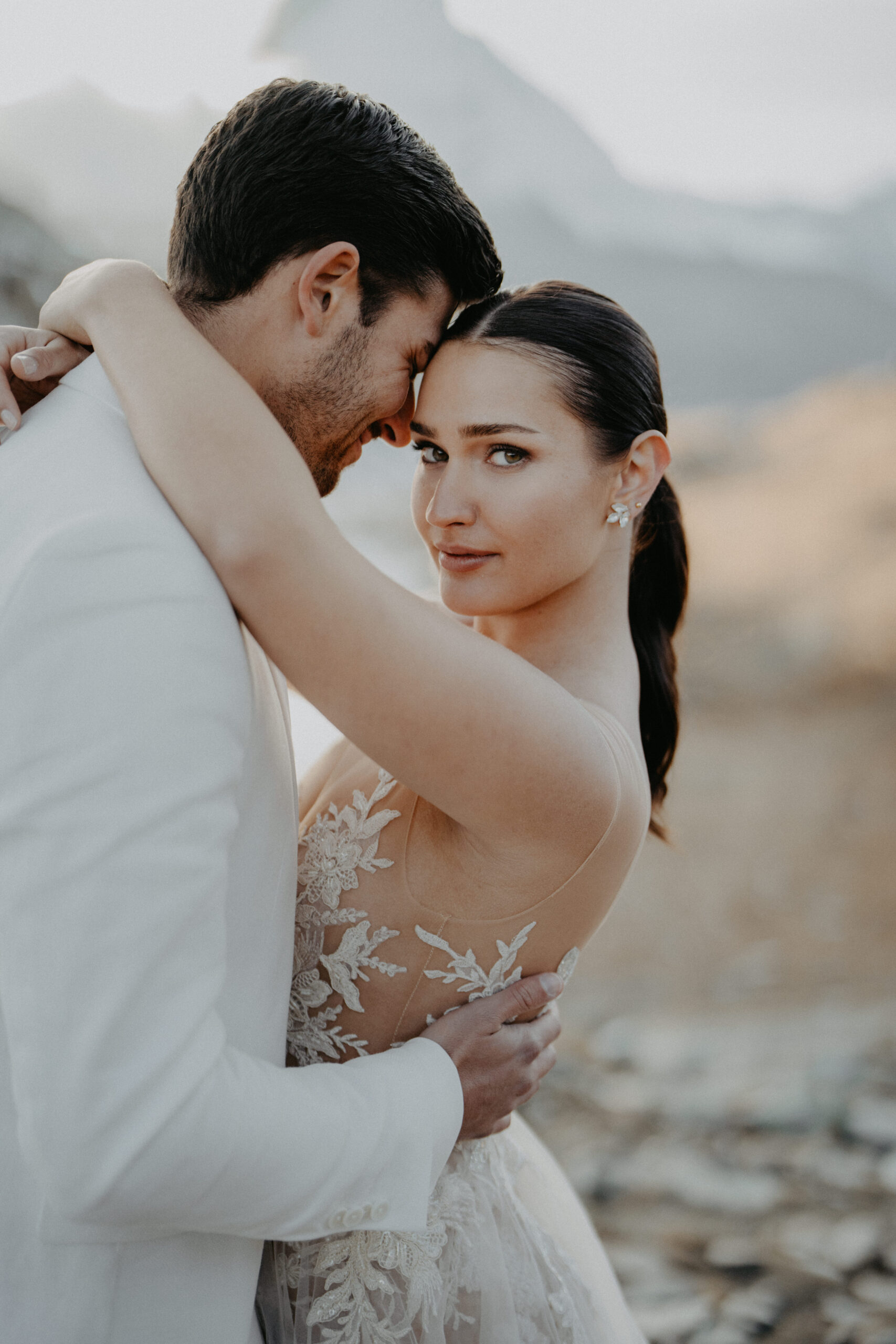 Die Braut im Spitzenkleid umarmt den Bräutigam im Freien in der Schweiz und blickt in die Kamera, während der Bräutigam seine Stirn an ihre schmiegt - ein perfekter Moment, eingefangen von einem Experten für Hochzeitsplanung.