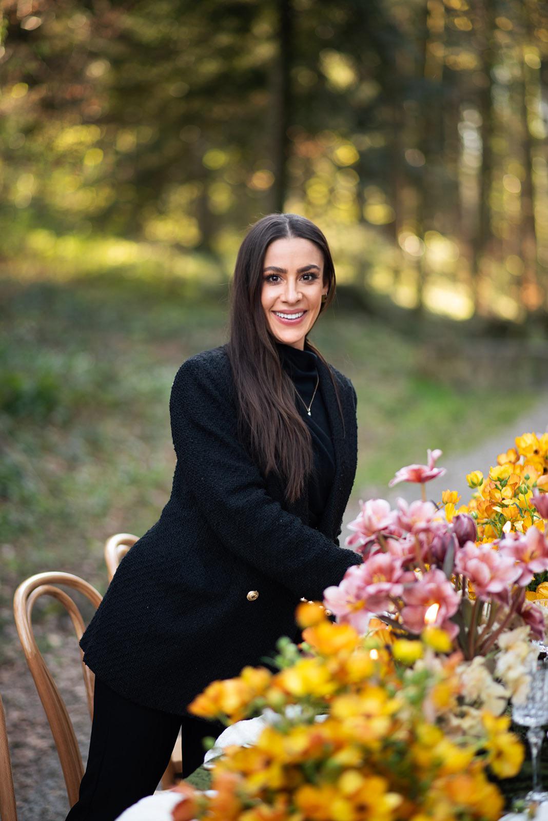 Frau mit langen braunen Haaren in einem schwarzen Outfit steht an einem Tisch mit bunten Blumen im Freien und zeigt ihre Eleganz als Hochzeitsplaner in der Schweiz.