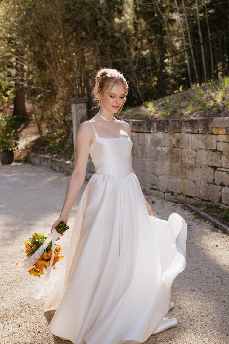 Eine Braut in einem weißen Kleid hält einen orangefarbenen Blumenstrauß und geht an einem sonnigen Tag im Freien spazieren, geführt von ihrem Hochzeitsplaner für eine perfekte italienische Feier.