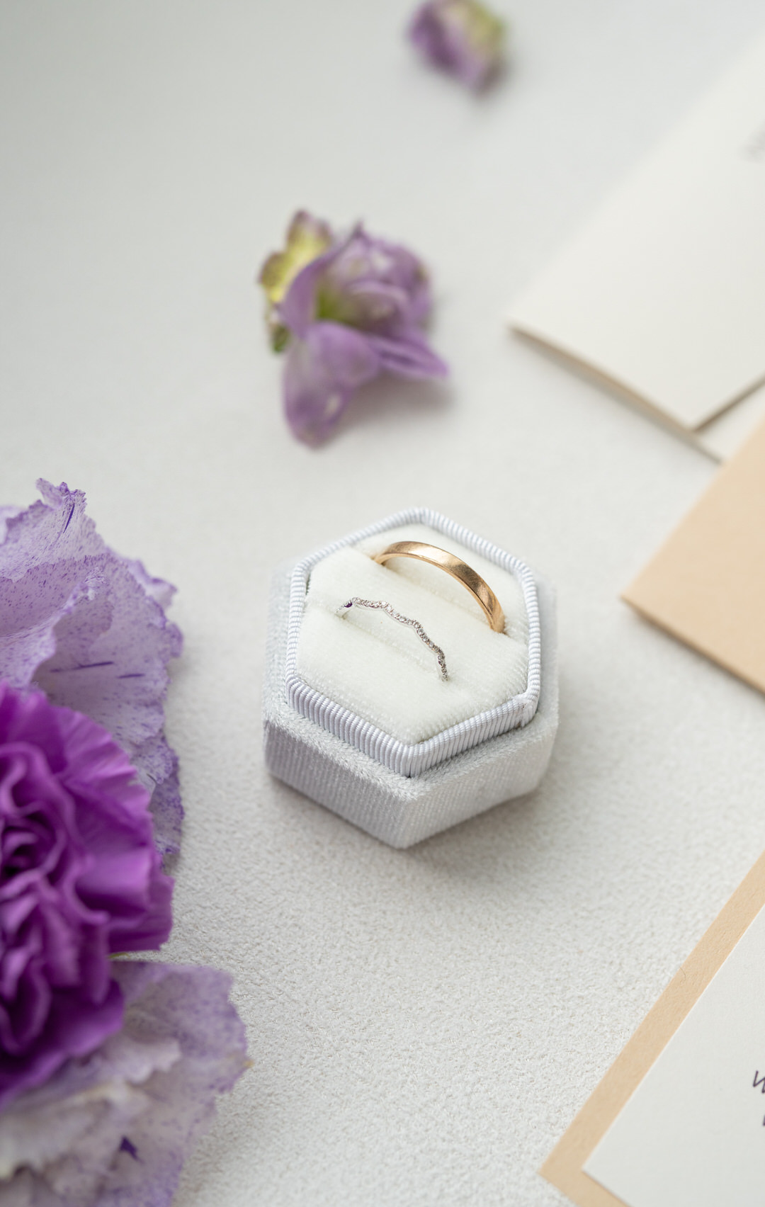 Goldene Eheringe in einer sechseckigen weißen Schachtel, umgeben von lila Blumen und Umschlägen - die perfekte Inspiration für jeden Hochzeitsplaner oder diejenigen, die ihre Hochzeitsplanung beginnen.