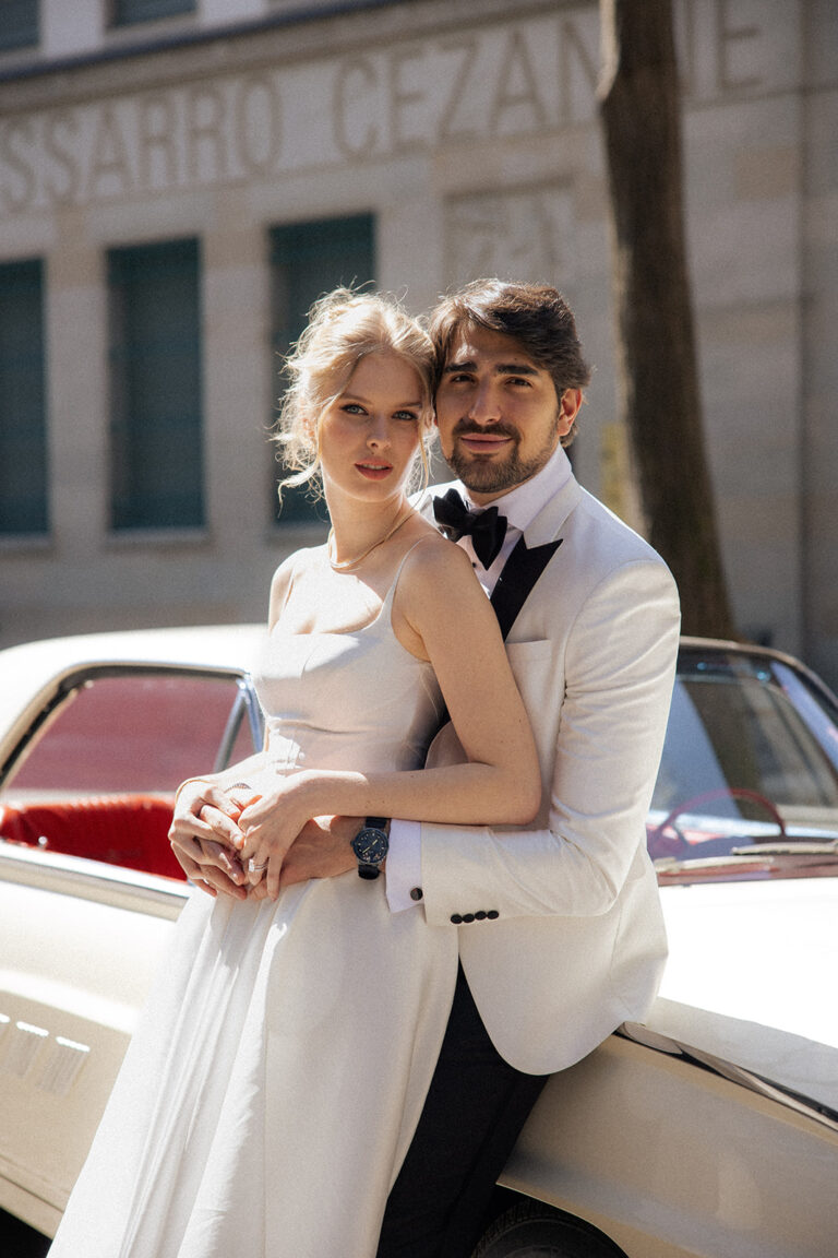 Ein formell gekleidetes Paar posiert an einem sonnigen Tag neben einem Oldtimer und fängt so die Eleganz ihres besonderen Moments in der Schweiz ein, perfekt arrangiert von ihrem Hochzeitsplaner.