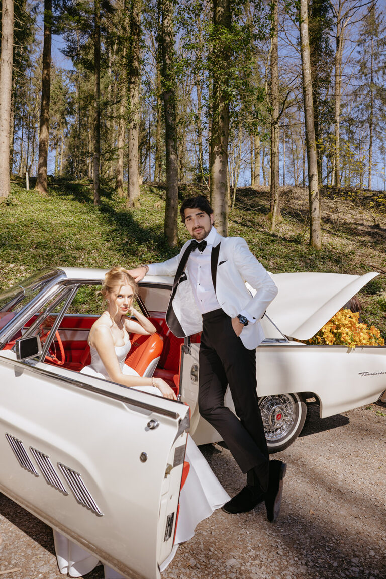 Eine Braut sitzt in einem weißen Oldtimer, während der Bräutigam neben ihr in einer bewaldeten Umgebung im Freien steht und den Zauber der Hochzeitsplanung in der Schweiz einfängt.