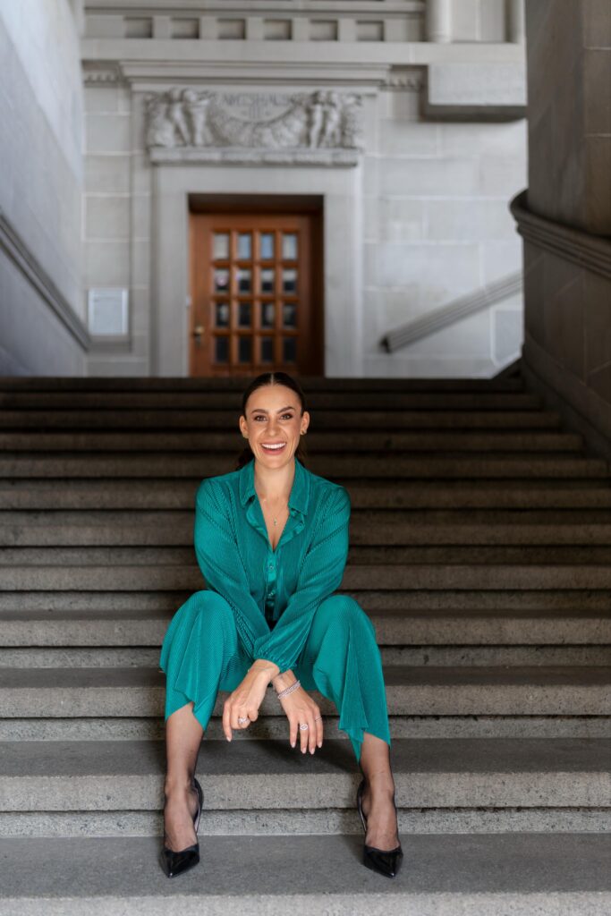 Eine Frau in einem blaugrünen Outfit sitzt auf einer Steintreppe in einem Gebäude mit klassischer Architektur und lächelt in die Kamera - vielleicht träumt sie von einer eleganten Hochzeitsplanung in Italien.