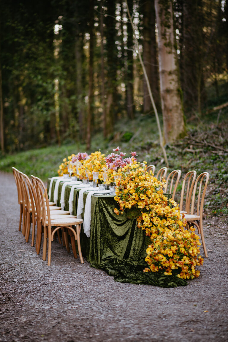 Ein Tisch im Freien mit grünem Samttuch und überquellenden gelben Blumenarrangements, umgeben von Holzstühlen - perfekt für einen Wedding Planner, der eine elegante, von Italien inspirierte Feier plant.