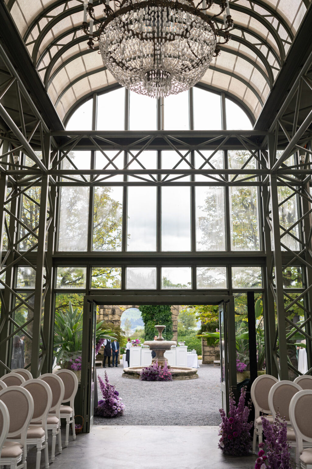 Home 39 Elegantes gläsernes Atrium für eine Veranstaltung, mit weißen Stühlen und lila Blumenarrangements, die nach draußen führen - perfekt für Paare, die einen raffinierten schweizerischen oder italienischen Touch von einem erfahrenen Wedding Planner suchen.