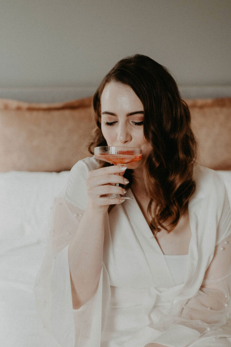 Eine Frau in einem weißen Gewand sitzt auf einem Bett und nippt lächelnd an einem Getränk aus einem Coupe-Glas - die perfekte Inspiration für jeden Hochzeitsplaner, der entspannte Brautmomente sucht.
