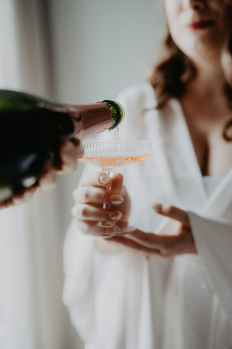 Eine Person in Weiß hält ein Glas in der Hand, in das jemand Sekt einschenkt - ein fröhlicher Moment, inspiriert von der Hochzeitsplanung in Italien.