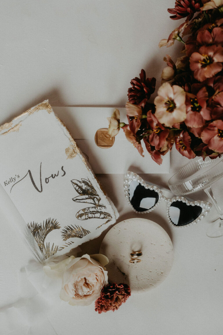 Ein flaches Layout von einer Broschüre mit dem Eheversprechen, einer Sonnenbrille, Blumen, Eheringen und einem Umschlag auf einer weißen Fläche - die perfekte Inspiration für Ihre Hochzeitsplanung mit einem Wedding Planner in der Schweiz.