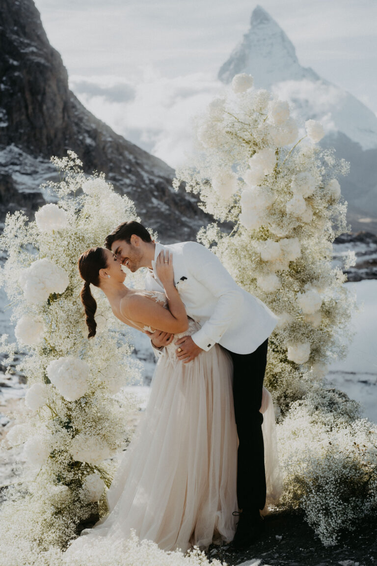 Eine Braut und ein Bräutigam geben sich einen romantischen Kuss in den Schweizer Bergen, umgeben von weißen Blumen - ein unvergesslicher Moment, den jeder Hochzeitsplaner gerne gestalten würde.