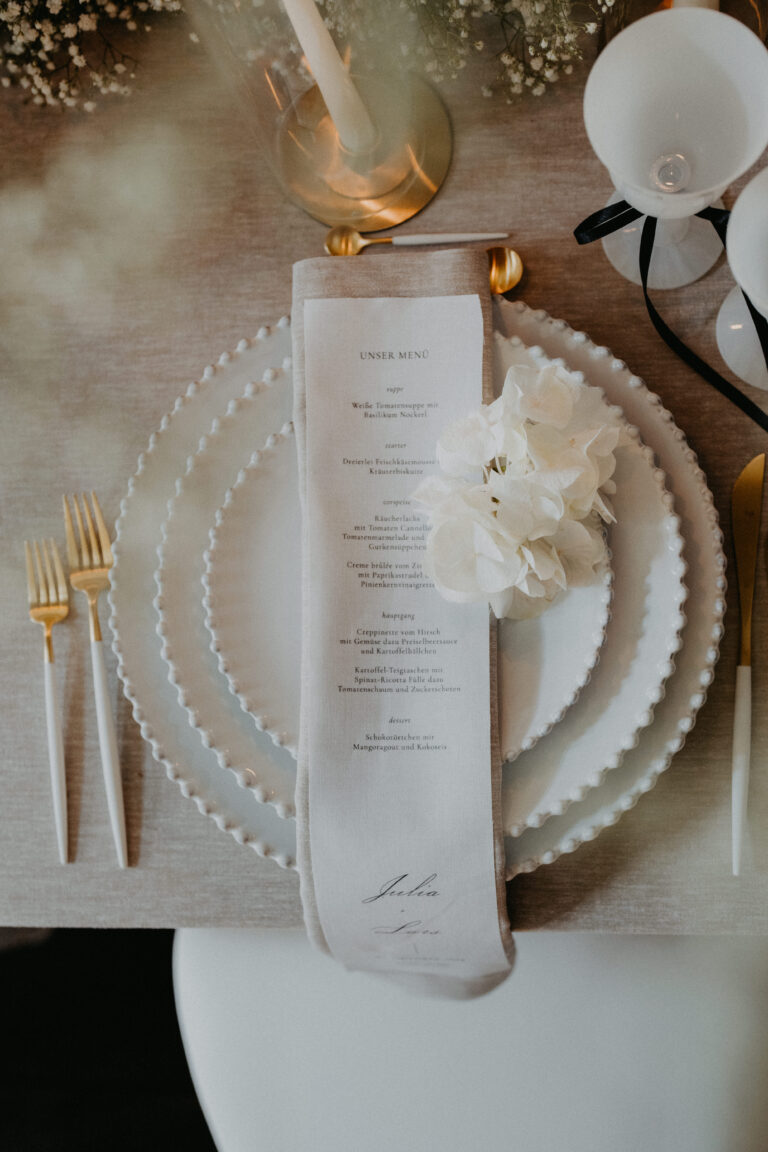 Elegant gedeckter Tisch mit weißen Tellern, goldenem Besteck, einer Menü-Serviette und einer weißen Blumendekoration - die perfekte Inspiration für Hochzeitsplaner in der Schweiz oder Italien.
