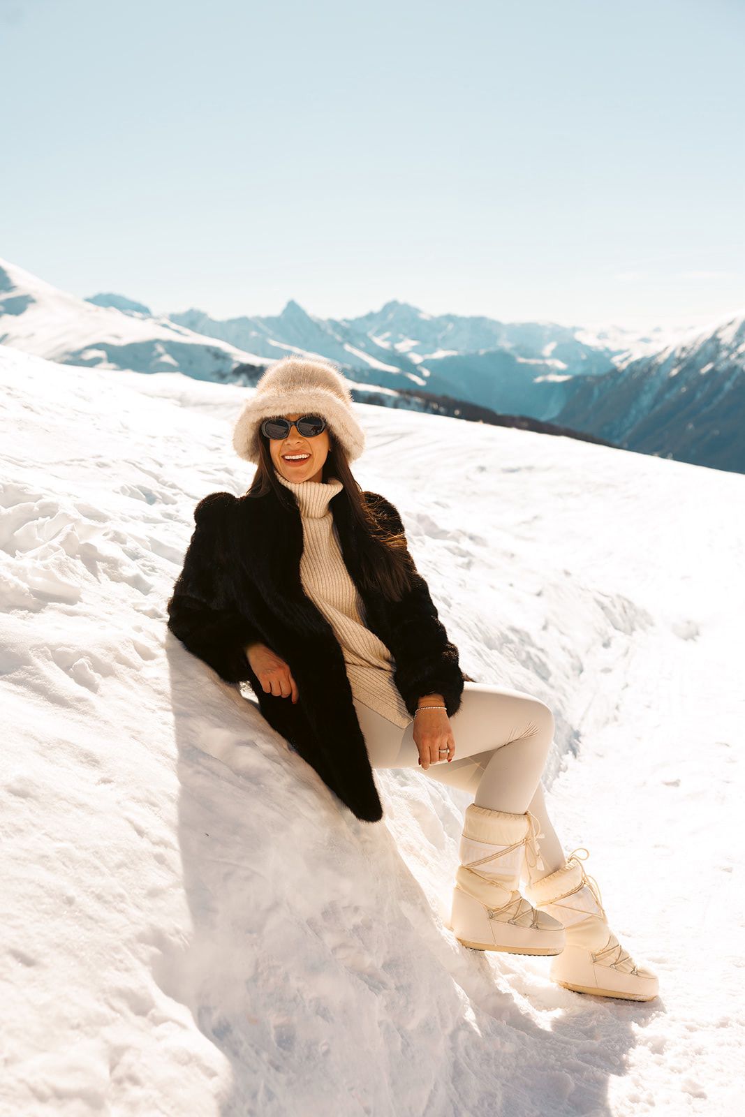 Frau in Winterkleidung lächelt und sitzt auf einem verschneiten Hang mit majestätischen Schweizer Bergen im Hintergrund unter klarem Himmel-perfekte Inspiration für winterliche Hochzeitsplanung.