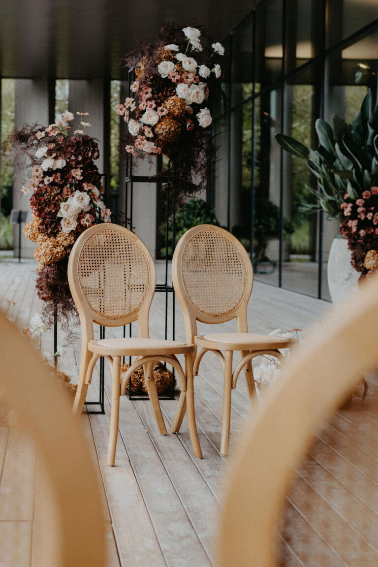 Zwei Holzstühle auf einer Terrasse in Italien, umgeben von Blumenarrangements und großen Fenstern - die perfekte Inspiration für Ihre Hochzeitsplanung.