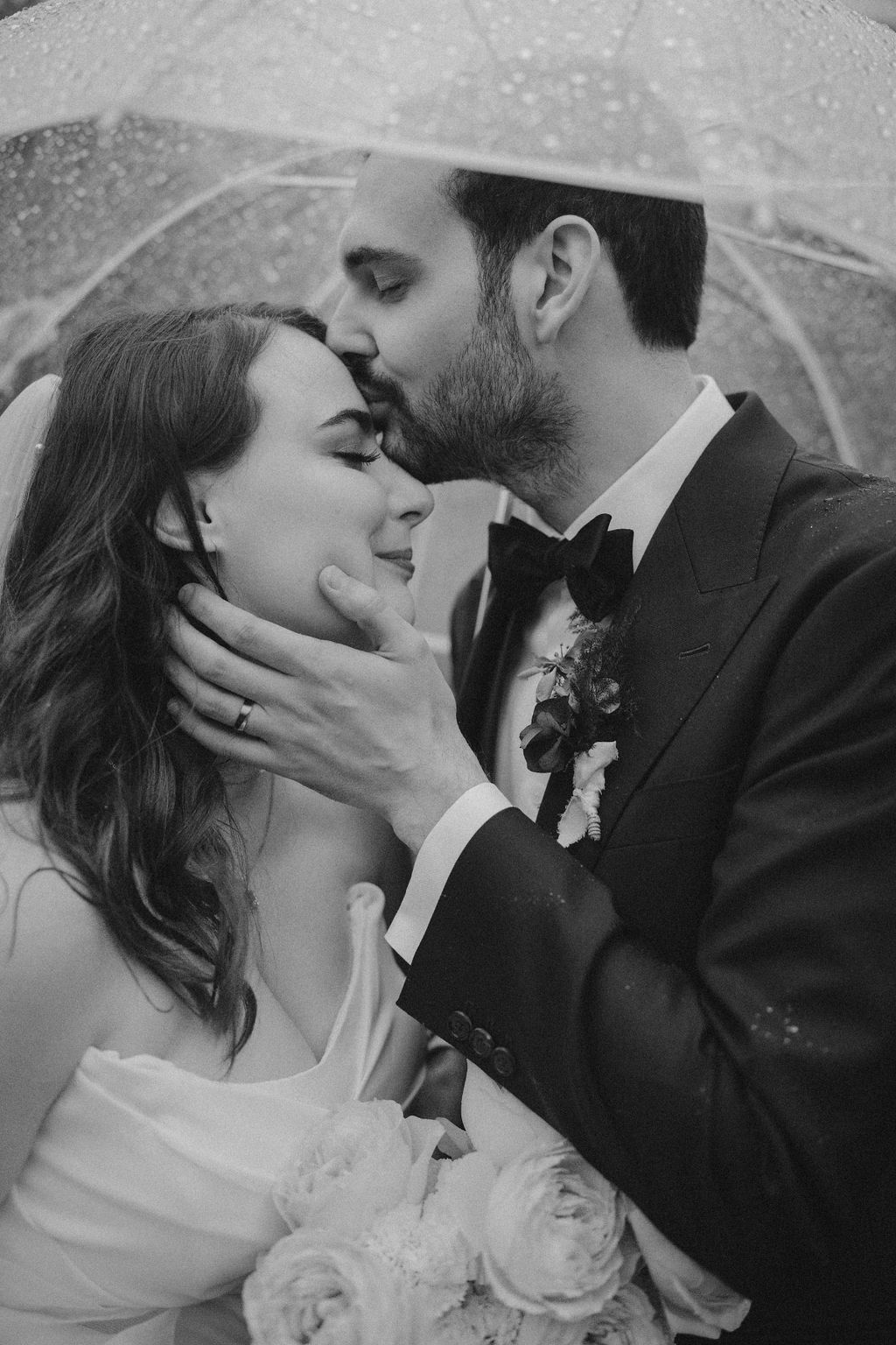 Home 45 Ein Bräutigam küsst seine Braut am Hochzeitstag unter einem durchsichtigen Regenschirm auf die Stirn und hält dabei einen Blumenstrauß.