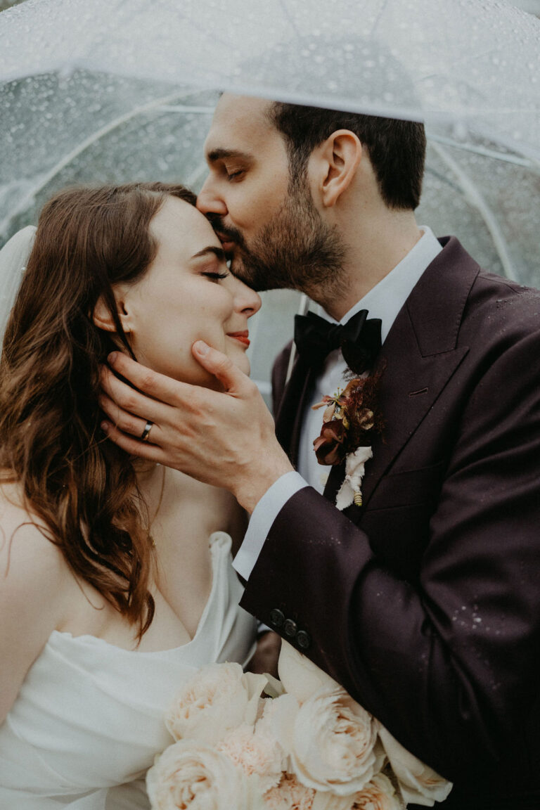 Der Bräutigam küsst die Stirn der Braut unter einem klaren Regenschirm in der Schweiz, beide halten weiße Rosen in der Hand und sehen glücklich und verliebt aus - ein perfekter Moment, eingefangen von einem erfahrenen Hochzeitsplaner.