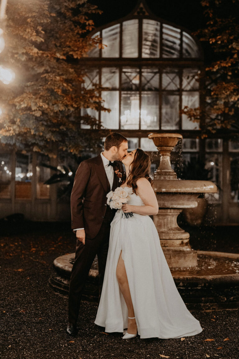 Eine Braut und ein Bräutigam küssen sich nachts vor einem Brunnen, gekleidet in Hochzeitskleidung - ein bezaubernder Moment, eingefangen von Ihrem Italien Wedding Planner für eine perfekte Hochzeitsplanung.