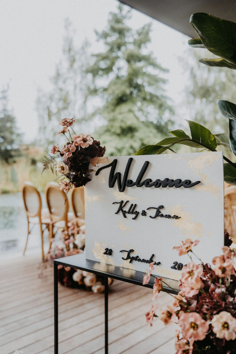 Ein Hochzeits-Willkommensschild mit Blumen an einer Holzterrasse, Stühlen und Bäumen im Hintergrund - perfekt für eine elegante Hochzeitsplanung in der Schweiz oder Italien.