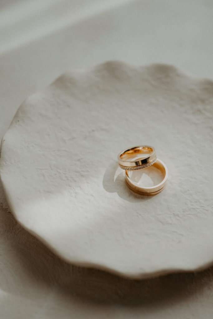 Zwei goldene Eheringe ruhen auf einer strukturierten, cremefarbenen Keramikplatte in sanftem natürlichem Licht - ein elegantes Detail, das jedem erfahrenen Wedding Planner gefallen würde.