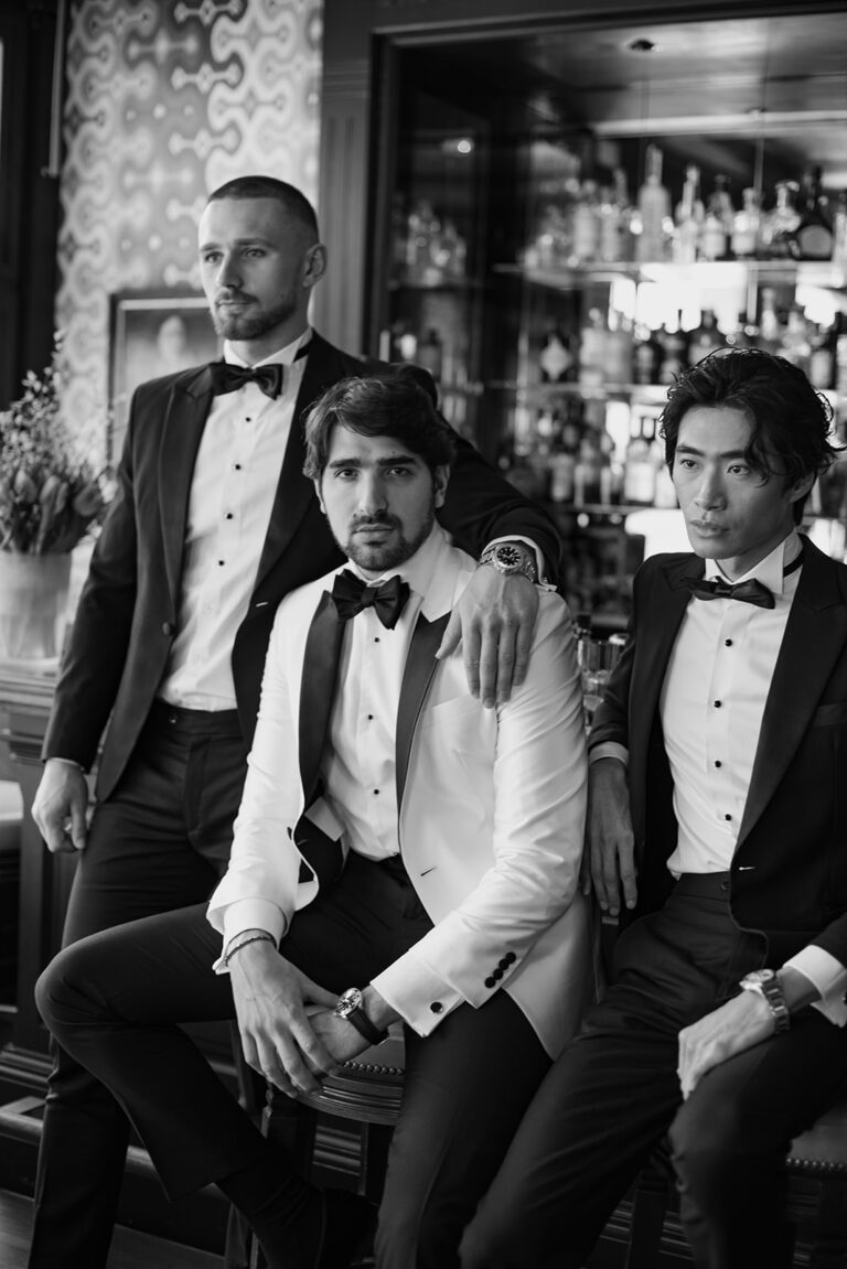 Drei Männer im Smoking posieren gemeinsam in einer stilvollen Bar - einer im weißen Jackett, zwei in Schwarz - und fangen die Eleganz eines Schweizer Hochzeitsplaners ein.