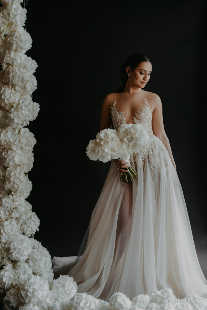 Eine Braut in einem durchsichtigen, eleganten Kleid hält weiße Blumen und steht neben einer Wand aus weißen Blüten vor einem dunklen Hintergrund - die perfekte Inspiration für Ihre Hochzeitsplanung mit Hilfe eines erfahrenen Wedding Planners.