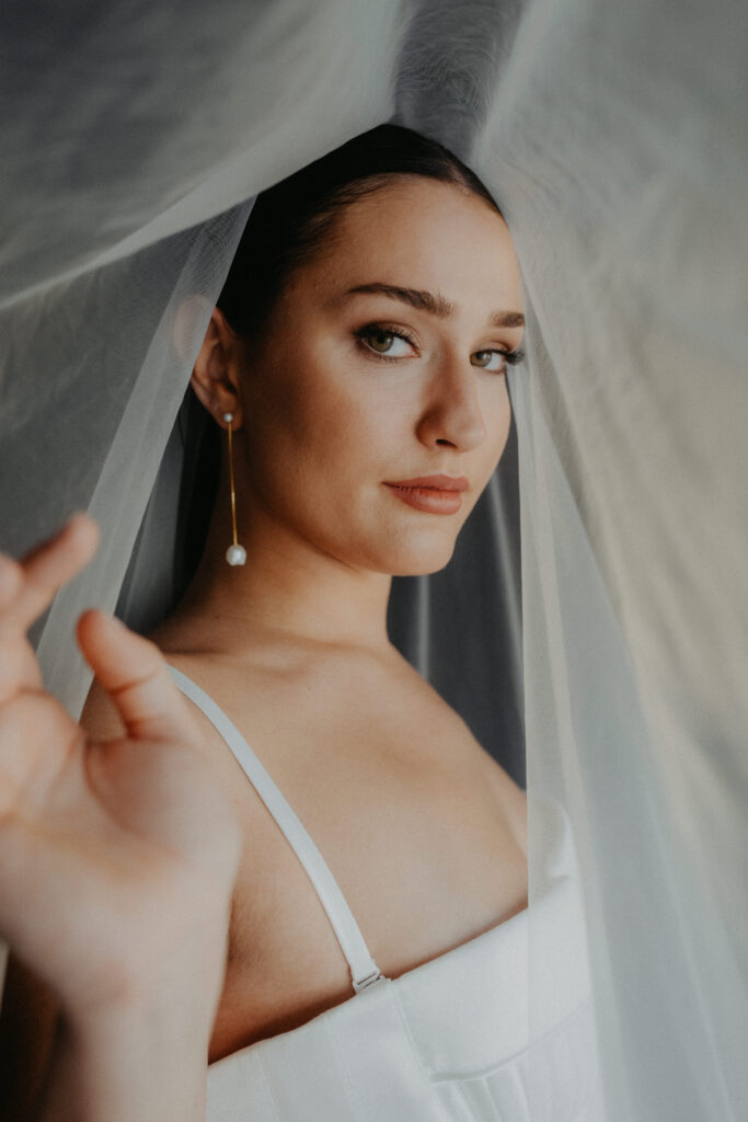 Eine Frau in einem weißen Kleid mit Schleier blickt in die Kamera und hält den durchsichtigen Stoff sanft an ihr Gesicht - ein eleganter Moment, der sich perfekt für die Hochzeitsplanung oder den Wedding Planner in Italien eignet.
