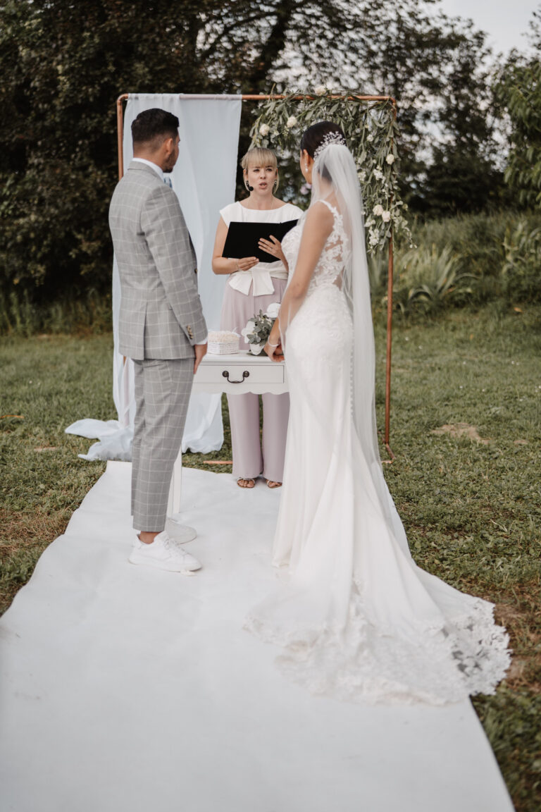 Die Braut und der Bräutigam stehen bei ihrer Hochzeitszeremonie, die von ihrem Hochzeitsplaner in der Schweiz wunderschön koordiniert wurde, vor einem Trauredner im Freien auf einem weißen Gang.
