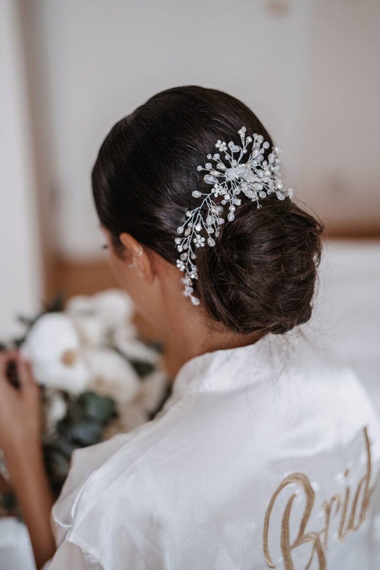 Frau mit dunklem Haar in einem niedrigen Dutt mit einem schmuckvollen Haarteil geschmückt, trägt ein weißes Gewand mit Braut auf dem Rücken-perfekt für Hochzeitsplanung in der Schweiz oder Italien.