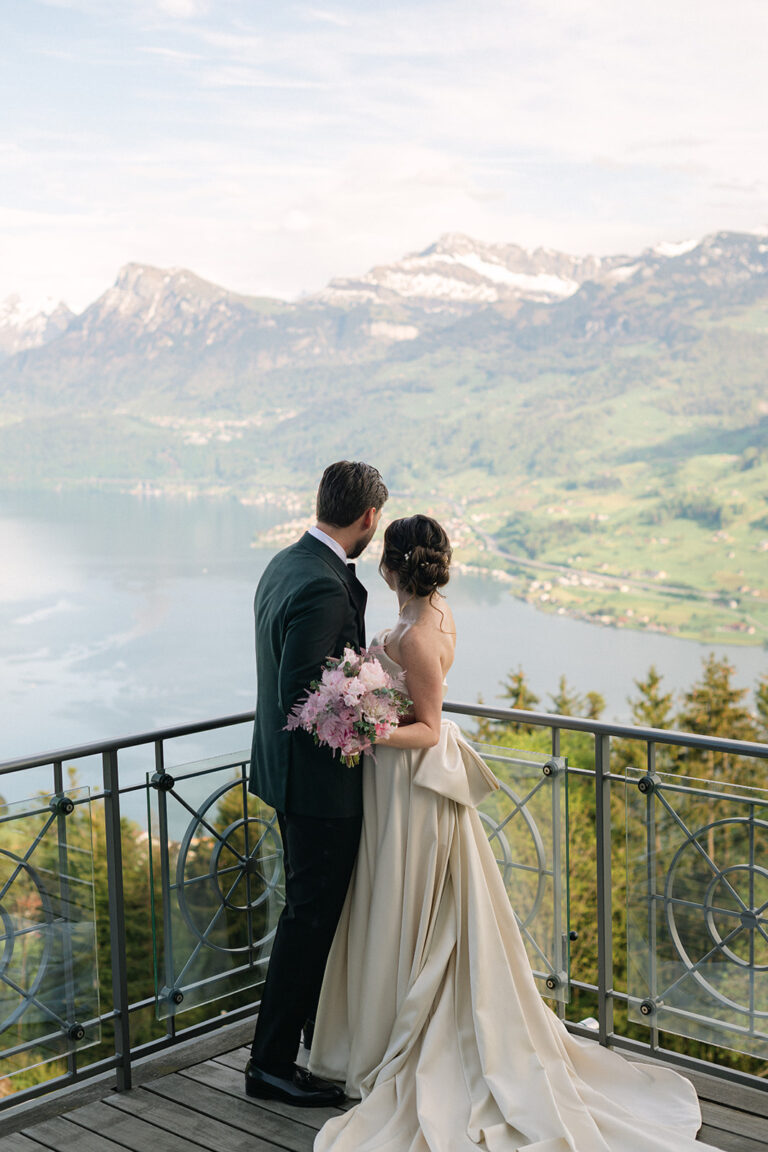 Eine Braut und ein Bräutigam stehen auf einem Balkon mit Blick auf einen See und die Berge in der Schweiz und halten einen rosafarbenen Blumenstrauß in der Hand - ein unvergesslicher Moment, der von ihrem Wedding Planner geschaffen wurde.