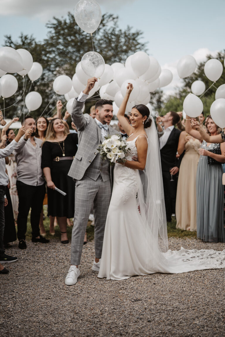 Braut und Bräutigam feiern im Freien, mit weißen Luftballons in der Hand, umgeben von jubelnden Gästen mit Luftballons - die perfekte Inspiration für Hochzeitsplanung in Italien oder der Schweiz.