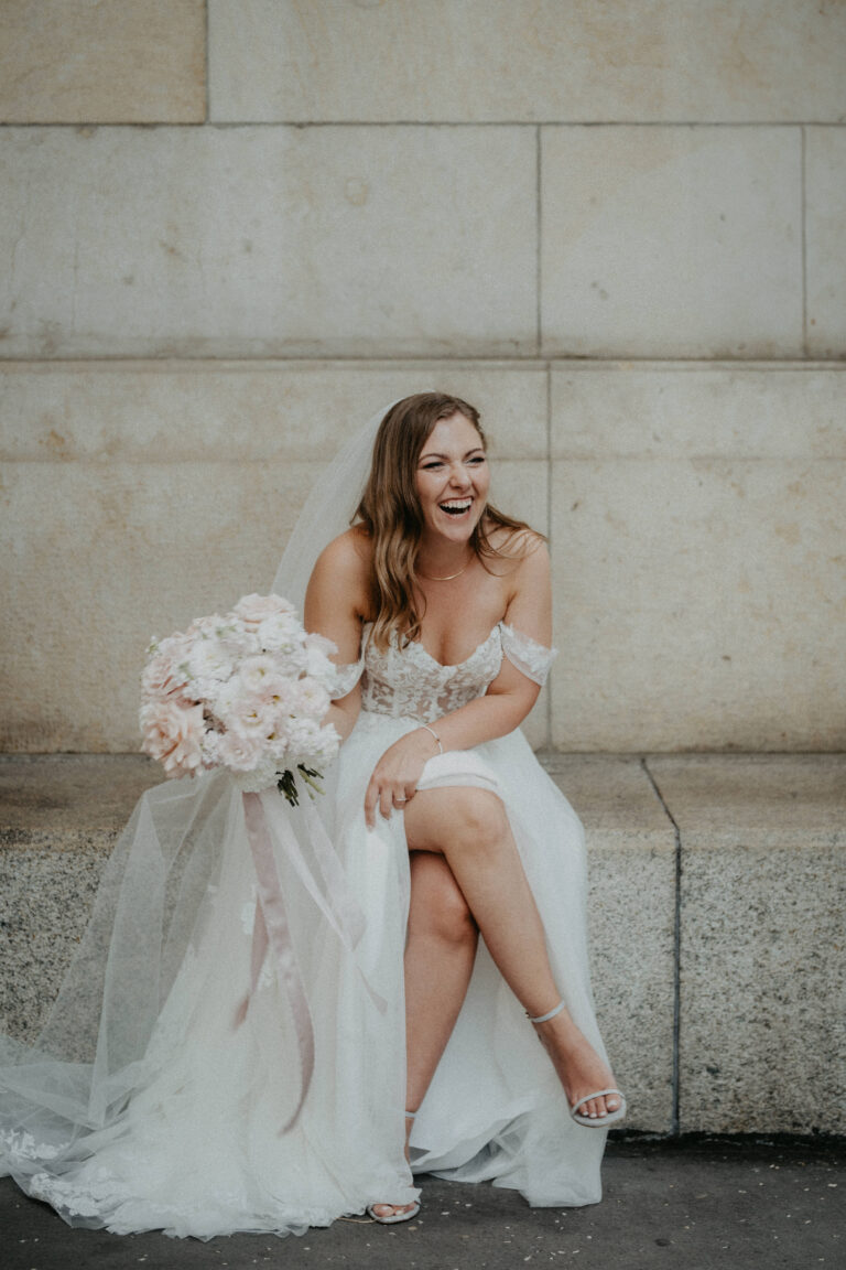 Eine lächelnde Braut in einem weißen Kleid mit einem Blumenstrauß in der Hand, die auf einer Steinbank vor einer beigen Wand sitzt - die perfekte Inspiration für jeden Wedding Planner in der Schweiz oder in Italien.