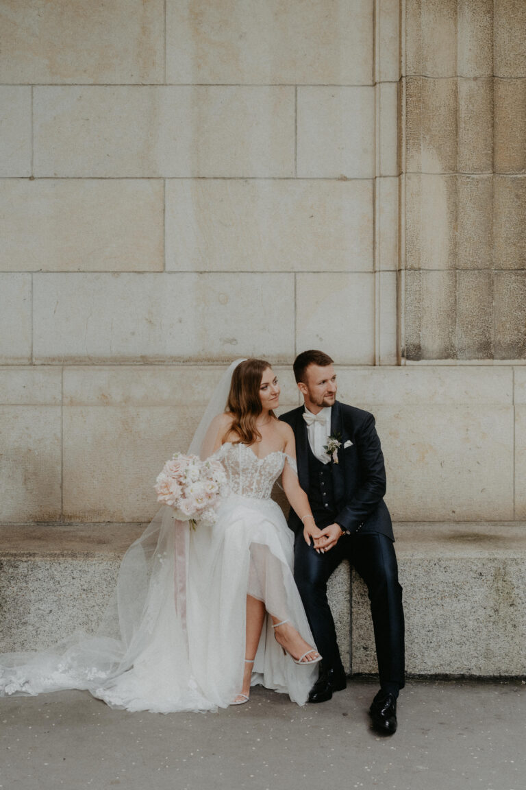Braut und Bräutigam sitzen zusammen auf einer Steinbank, halten sich an den Händen und schauen in eleganter Hochzeitskleidung in die Ferne - ein idyllischer italienischer Moment, kuratiert von einem erfahrenen Hochzeitsplaner.