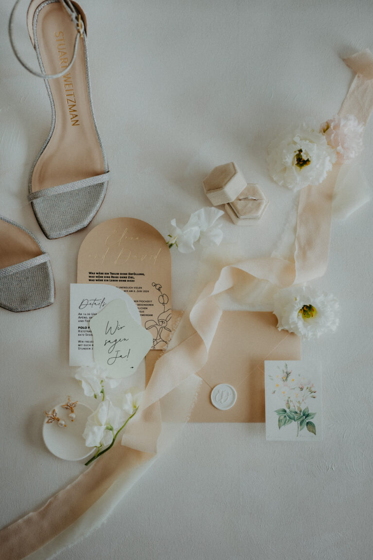 Flache Hochzeitseinladungen, weiße Blumen, Schleifenband, Ringe in einer Schachtel und ein Paar hochhackige Sandalen - die perfekte Inspiration für die Hochzeitsplanung, egal ob Ihr Traumfest in der Schweiz oder in Italien stattfindet.