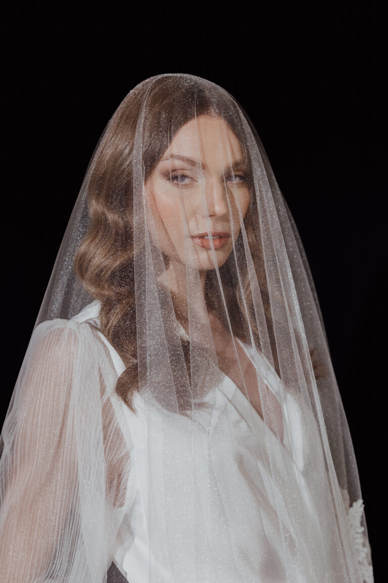 Eine Frau in einem weißen Kleid mit gewelltem Haar, die einen durchsichtigen, glitzernden Schleier trägt, steht vor einem dunklen Hintergrund - die perfekte Inspiration für jeden italienischen Hochzeitsplaner.