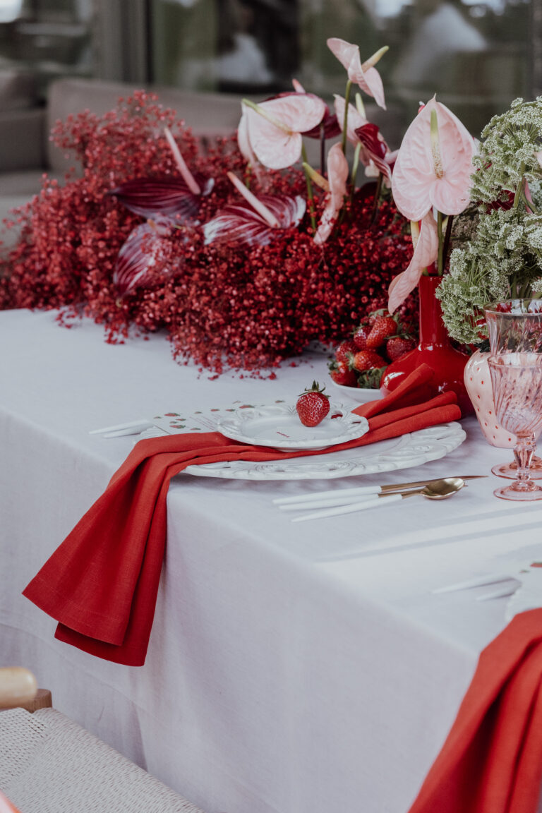 Elegant gedeckter Tisch mit roten Servietten, rosa Blumen und einer einzelnen Erdbeere auf einem Teller - die perfekte Inspiration für alle Hochzeitsplaner in der Schweiz, die auf der Suche nach romantischen Akzenten sind.