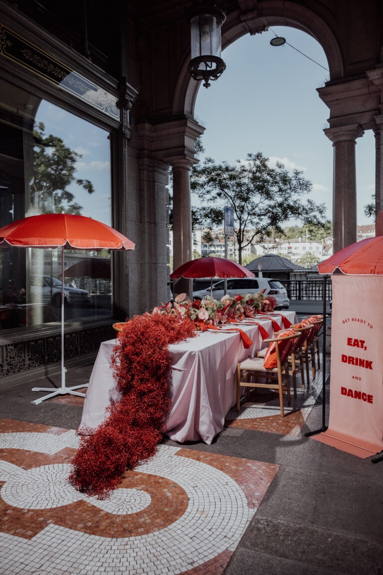Ein Tisch im Freien mit roter Dekoration, Blumenschmuck, roten Sonnenschirmen und einem Schild mit der Aufschrift "Essen, Trinken und Tanzen" - die perfekte Inspiration für jeden Hochzeitsplaner in Italien oder Hochzeitsplaner, der elegante Ideen für seine Feier sucht.