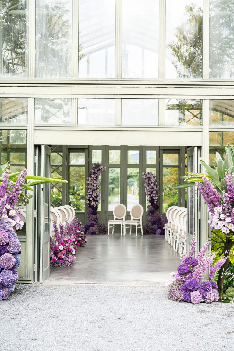 Ein gläserner Wintergarten für eine Hochzeit mit weißen Stühlen und violetten Blumenarrangements, die den Gang säumen - eine elegante Kulisse, die von einem professionellen Hochzeitsplaner gestaltet wurde.