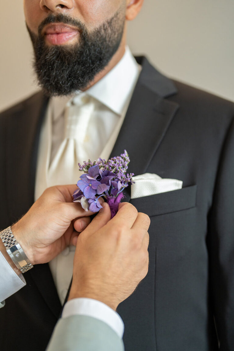 Eine Person steckt eine lilafarbene Boutonniere an die schwarze Anzugsjacke eines Mannes mit weißer Krawatte und Einstecktuch und zeigt damit die elegante Hochzeitsplanung durch einen erfahrenen Wedding Planner.