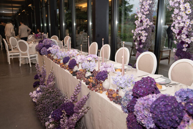 Eleganter Banketttisch mit weißen Stühlen, dekoriert mit lila Blumen, Kerzen und Obst - die perfekte Inspiration für die Hochzeitsplanung in der Schweiz oder Italien.