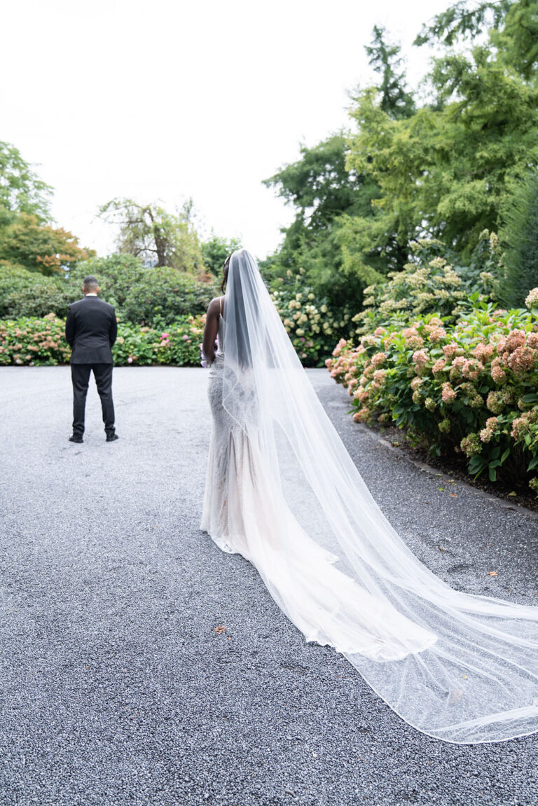 Eine Braut mit langem Schleier geht auf einen Bräutigam zu, der auf einem von Grün umgebenen Kiesweg steht. Ihre Schweizer Hochzeit wurde von einem erfahrenen Hochzeitsplaner zum Leben erweckt.