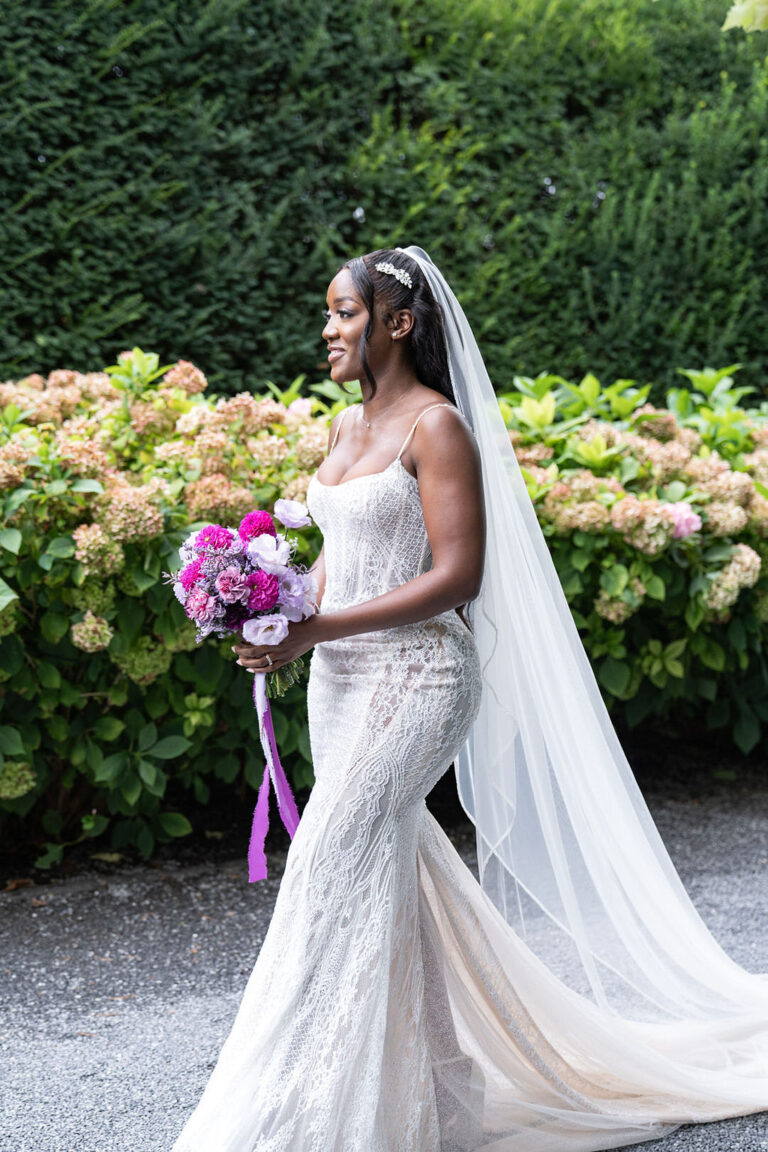 Eine Braut in Spitzenkleid und Schleier hält einen Strauss aus rosa und lila Blumen und geht im Freien spazieren - die perfekte Inspiration für Ihre Hochzeitsplanung mit einem erfahrenen Wedding Planner in der Schweiz.