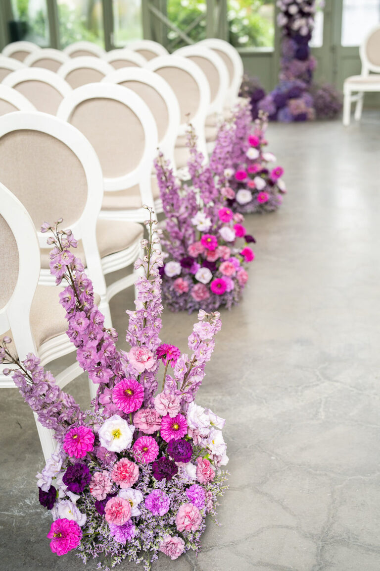 Weiße Stuhlreihen mit rosafarbenen und violetten Blumenarrangements säumen den Gang in einer hellen Halle, wunderschön arrangiert von einem erfahrenen Hochzeitsplaner.
