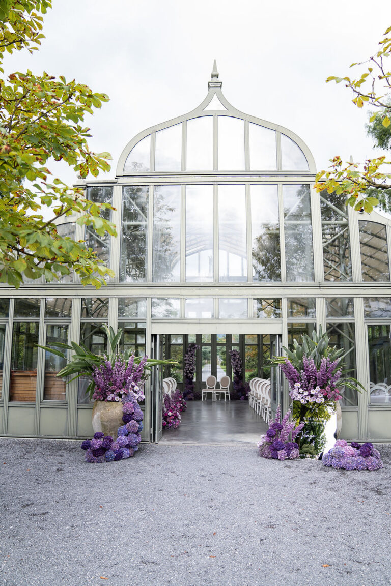Ein gläsernes Gewächshaus mit offenen Türen, weißen Stühlen im Inneren und lilafarbenen Blumenarrangements am Eingang - perfekt für Ihre italienische Hochzeitsplanung unter Anleitung eines erfahrenen Wedding Planners.