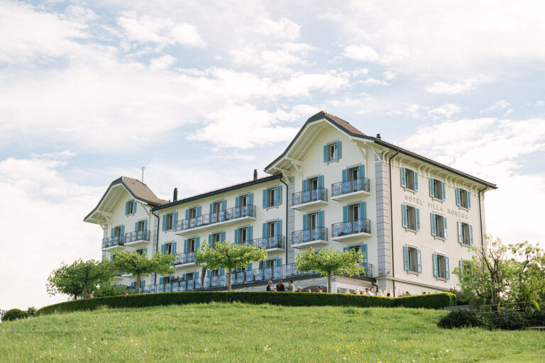 Das elegante weiße Hotel mit Balkonen liegt auf einem grasbewachsenen Hügel unter einem teilweise bewölkten blauen Himmel.
