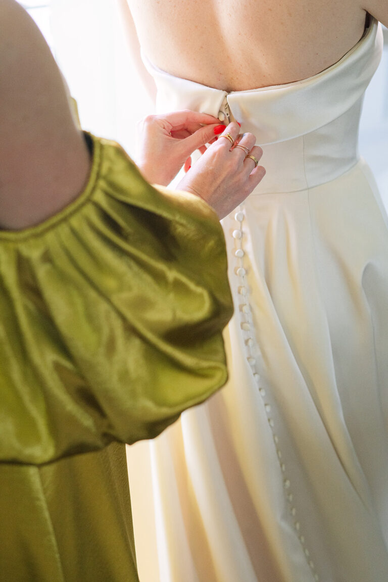 Ein Hochzeitsplaner in der Schweiz befestigt Knöpfe am Rücken des weissen Kleides einer Braut; eine andere Person trägt ein leuchtend grünes Outfit.
