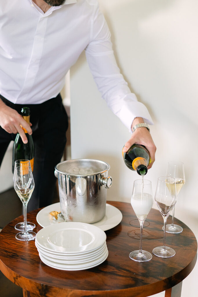 Eine Person gießt Champagner in Flöten neben einem Eimer mit Eis und gestapelten Tellern auf einem Holztisch ein - perfekt für eine elegante Hochzeitsplanung in der Schweiz.