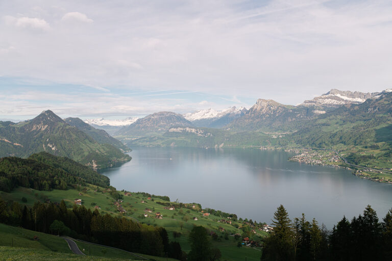 Ein ruhiger See, umgeben von grünen Hügeln, verstreuten Häusern und schneebedeckten Bergen unter einem wolkenverhangenen Himmel - die perfekte Inspiration für jeden Hochzeitsplaner, der eine atemberaubende Hochzeitslocation sucht.