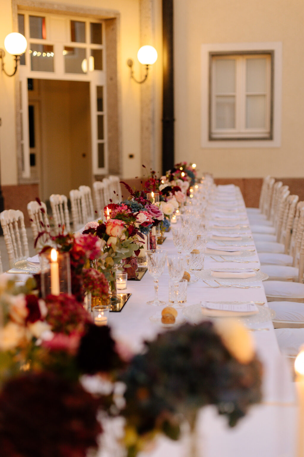 Home 24 Ein eleganter Esstisch im Freien mit Blumen, Kerzen und weißen Stühlen vor einem gelben Gebäude - perfekt für einen Schweizer Hochzeitsplaner, der eine romantische, von Italien inspirierte Feier plant.