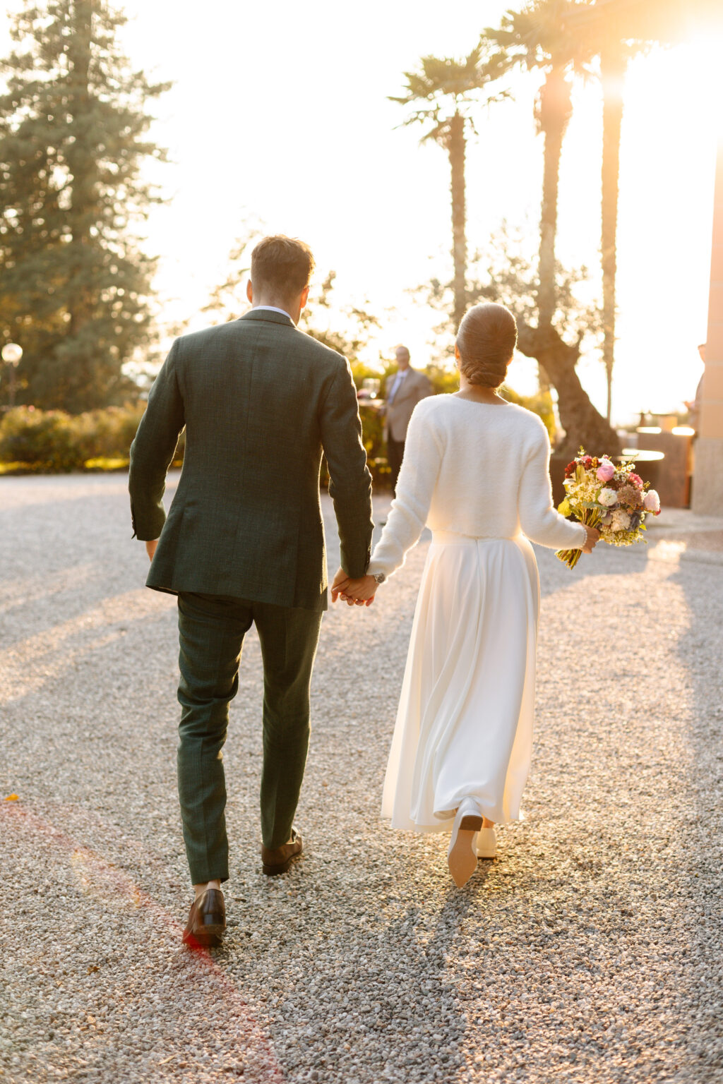 Home 21 Ein händchenhaltendes Paar, das bei Sonnenuntergang in Italien spazieren geht; die Frau hält einen Blumenstrauß - eine bezaubernde Szene für jeden Hochzeitsplaner.